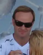 Photo de Barrichello, lunettes noires, signant un autographe à une femme blonde, vue de dos