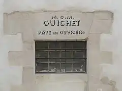 Fenêtre étroite percée dans un mur, sur lequel est inscrit « M.O.M. Guichet Paye des ouvriers ».