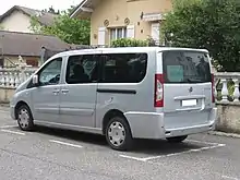 Photographie en couleurs d’une voiture grande capacité grise, garée devant une maison.