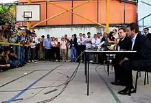 Deux hommes en costume, assis à une table, sur un terrain de basket, devant une foule de journalistes.