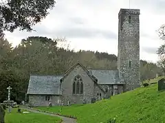 Photo d'un bâtiment allongé aux murs gris et au toit en ardoises, flanqué d'une très haute tour carrée sur la droite, à demi caché par un talus herbeux.