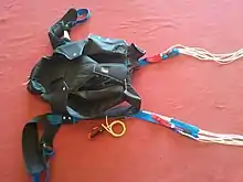 Le sac harnais avec les élévateurs de la voile de secours en bleu et ses commandes en rouge. La voile de secours se trouve en haut du sac harnais et la voile principale en bas.La poignée rouge et noire avec le jonc jaune c'est la poignée de libération qui libère la voile principale.Le seul élément manquant est la poignée d'ouverture du secours qui a été perdue : la récupération des poignées n'est pas une priorité quand on fait une PDS. Seule la sécurité est essentielle.