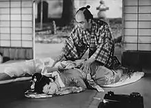 Une femme en kimono est alongée sur le tatami d'une pièce traditionnelle japonaise dont la porte coulissante s'ouvre sur un jardin. Un homme à genou tend les mains vers elle (image en noir et blanc).