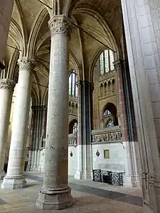 Au premier plan, des piliers, dont certains sont peints liés par des tirants de bois. En arrière-plan, un mur massif orné de bas-reliefs.