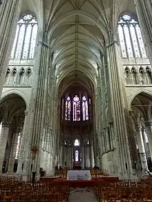Intérieur d'une église avec des voûtes très hautes soutenues par des piliers en pierre. En haut de l'image, grandes verrières colorées. En bas de l'image, 5 ouvertures délimitées par 6 gros piliers donnent accès à des chapelles.