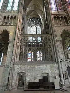 Derrière un mur massif en pierre, vue d'une partie d'une façade surmontée de verrières et d'une rosace en forme de fleur.