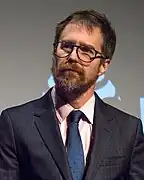 Photo en buste d'un homme barbu avec des lunettes et habillé en costume.