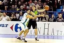 Homme de face, tenant le ballon dans sa main gauche, un adversaire, collé à lui, sur sur côté droit.