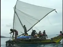 Photographie en couleur d'une pirogue à balancier sur laquelle se trouvent six hommes qui manœuvrent l'embarcation