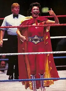 La catcheuse Sherri Martel portant une combinaison rouge, une cape rouge et jaune et une ceinture de championne de catch.