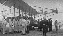 Photographie noir et blanc d'un avion, au sol, avec des matelots autour et un homme, en manteau sombre, qui marche vers la caméra.