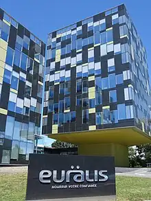 Photo d'un bâtiment moderne de plusieurs étages vitrés, au premier plan une plaque indiquant le nom de l'entreprise.