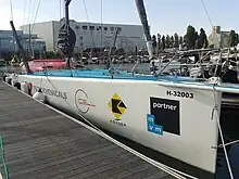 Voilier blanc amarré au ponton, vu de trois quarts avant. Pont bleu, rouf rouge.