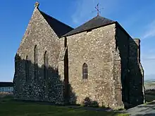 Photo d'un bâtiment aux murs gris avec des fenêtres hautes et étroites. Son toit en ardoises est surmonté d'une croix celtique en pierre d'un côté et d'une girouette de l'autre.