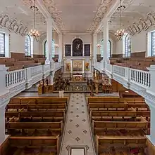 L'intérieur d'une grande salle aux murs blancs, un balcon de chaque côté. Des rangées de bancs en bois marron. Quelques reflets or au niveau du choeur.
