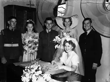 Une jeune femme en robe de mariée signe un registre de mariage en souriant. Derrière, le marié et quatre témoins en tenue militaire.