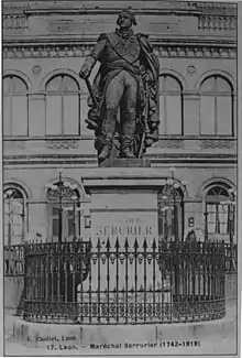 Photographie ancienne d'une statue en pied d'un maréchal de Napoléon en grand uniforme, tenant son sabre de la main gauche et son bâton de maréchal de la main droite. Autour du piédestal, une grille, et en arrière-plan, un bâtiment.