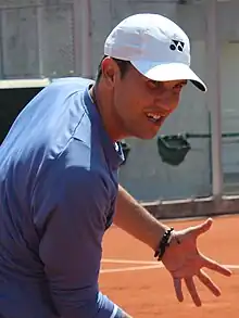 Sur un court en terre battue. Plan rapproché. Casquette blanche, survêtement bleu pétrole. Il explique quelque chose en mimant.