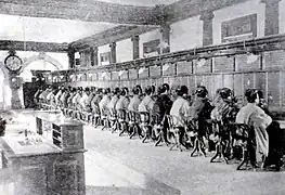 Photographie d'une très grande salle de travail ; un mur est intégralement occupé par des appareils téléphoniques ; une vingtaine de femmes assises et visibles de dos y travaillent.