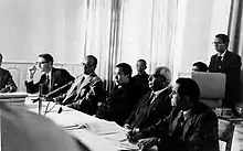 Photographie en noir et blanc montrant le dalaï-lama s'exprimant lors d'une conférence, entouré d'autres hommes.