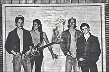 Photographie en noir et blanc de quatre hommes debout avec sur le mur un affiche de Marilyn Monroe