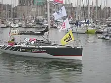 Un voilier noir et blanc avance au moteur dans un port. Sur la coque, on peut lire « Faber France » et « Dunkerque communauté urbaine ».