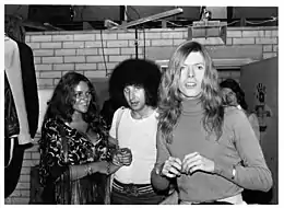 Photo en noir et blanc de trois personnes : une jeune femme brune au teint mat, un homme brun avec une coupe afro et un homme aux longs cheveux blonds ondulés qui regarde vers l'objectif