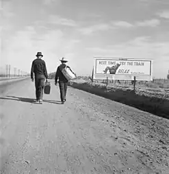 Deux hommes marchant le long de la route près d'un panneau d'affichage indiquant « La prochaine fois, essayez le train. Détendez-vous ». Mars 1937.