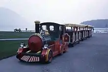Photographie en couleurs d’un petit train sur pneus multicolore avec trois wagons rouge et blancs sur une route au bord de l’herbe. En arrière-plan, on distingue un lac et des montagnes sous un ciel laiteux.
