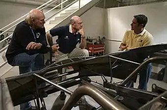 Trois hommes discutant devant une carlingue de voiture. Chacun a un pied posé sur le squelette de la voiture.