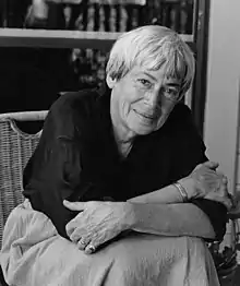 Photo portrait en noir et blanc d'Ursula Le Guin qui est assise et croise les bras devant une étagère de livres.
