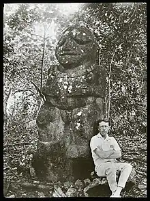 Un homme est assis contre une statue faisant deux fois sa taille. La statue stylisée représente un personnage ventru.