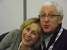 Une femme blonde adossée à un homme aux cheveux blancs portant des lunettes de vue, ils sont souriants.