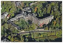 Photographie aérienne d'un paysage arboré montrant un grand bâtiment à plusieurs ailes, bodé au premier plan d'un ruisseau et d'une route.