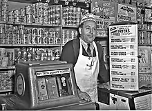 Photographie en noir et blanc d’un épicier souriant, cravaté, portant un tablier blanc à bretelle unique passant derrière le cou, coiffé d’un calot publicitaire ; l’homme s’appuie à une machine distributrice de café et à un comptoir surmonté de panneaux publicitaire ou annonçant les prix de marchandises. Derrière lui, l’étagère qui court tout le long du mur est remplie d’une multitude de boîtes de conserve, parfois cachées par des calicots publicitaires supplémentaires, donnant l’impression qu’il n’y a plus 1 cm2 de disponible.