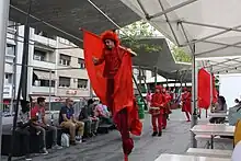 Parade rouge comprenant des échassiers, des danseurs et des musiciens percussionnistes.